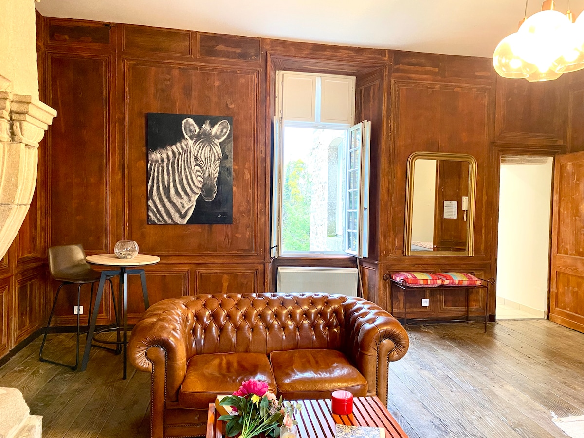 The living area features warm wooden paneling and a large window offering a view of the greenery outside. A dark leather sofa is positioned near a small round table with glassware on it. A vibrant artwork of a zebra adds a touch of character to the walls.