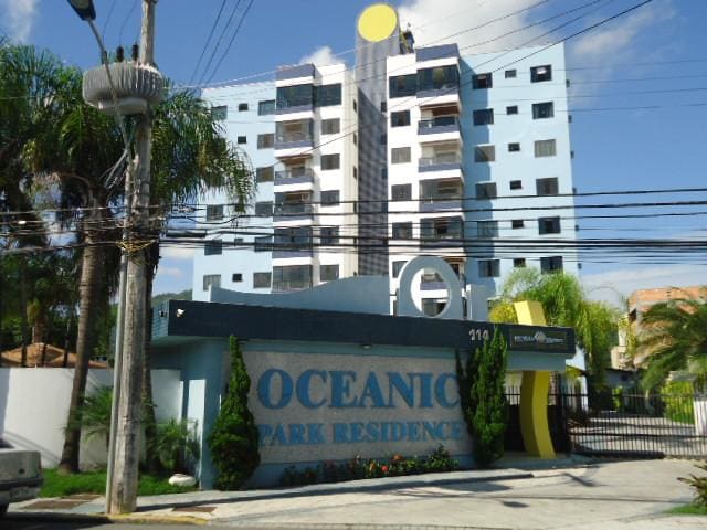 The exterior of the Oceanic Park Residence is presented, showcasing a multi-story building with a blue facade. The entrance is marked by a prominent sign, surrounded by greenery. Power lines are visible overhead, complementing the urban landscape.