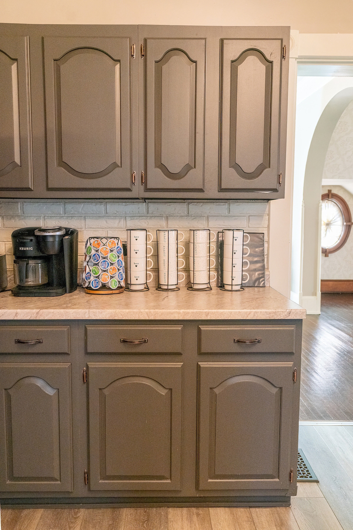 A well-organized kitchen countertop features gray cabinets with raised panel doors. An array of white containers lines the back, complemented by a colorful coffee maker. Lightly textured flooring extends into a visible hallway, connecting to an archway that hints at additional spaces within the home.
