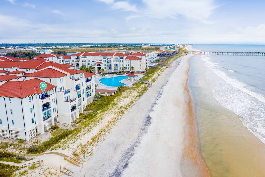 An aerial view captures the beachfront property, showcasing a long stretch of sandy shoreline meeting the ocean. The villa's distinctive architecture is visible, with red roofs and balconies overlooking the water. The nearby pool area adds to the resort's appeal, surrounded by lush greenery.