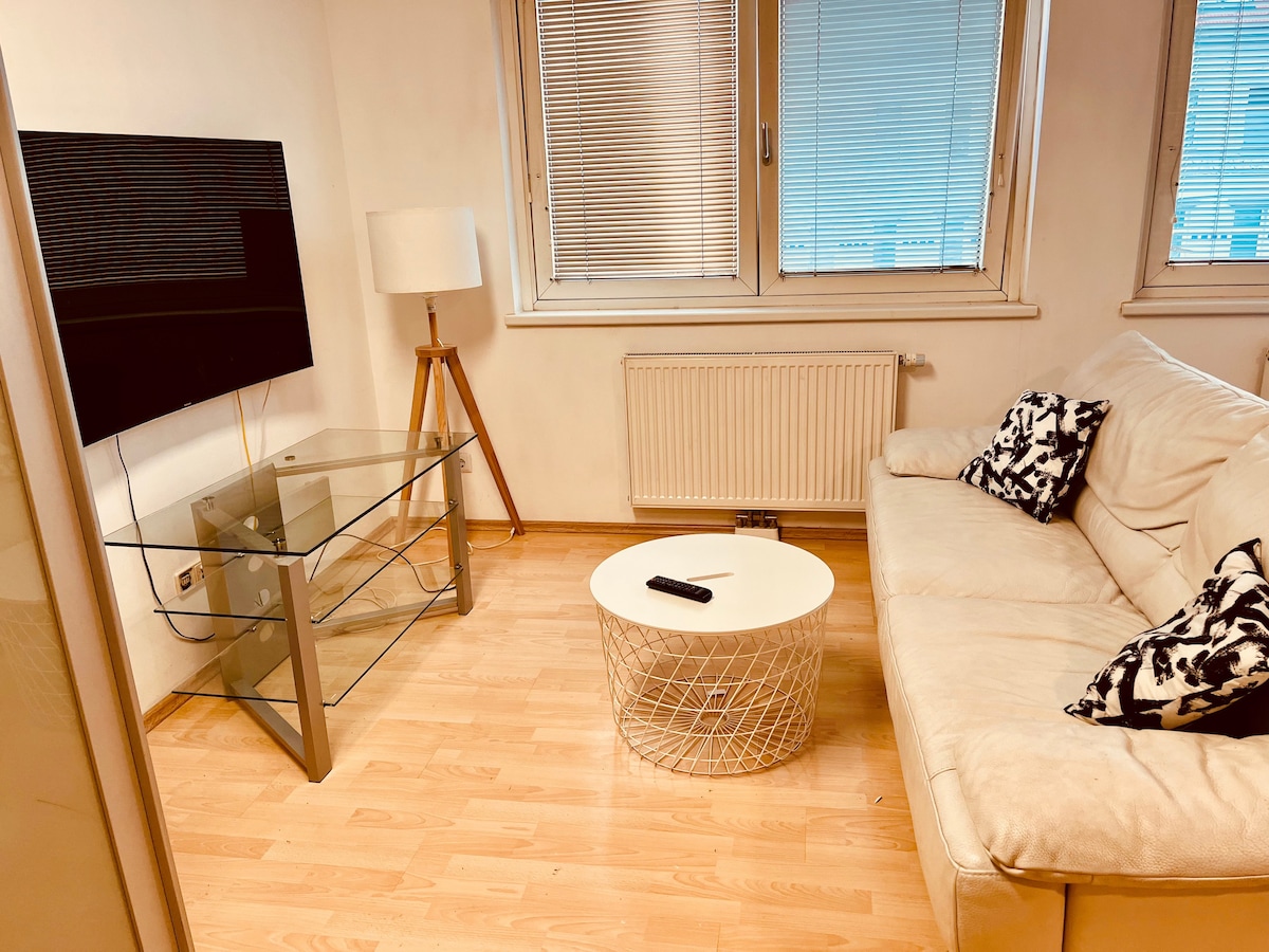 A living area is presented with a light-colored sofa and a circular coffee table. A flat-screen television is mounted on the wall opposite a glass TV stand. Natural light filters through the horizontal window blinds, illuminating the warm wooden floor.
