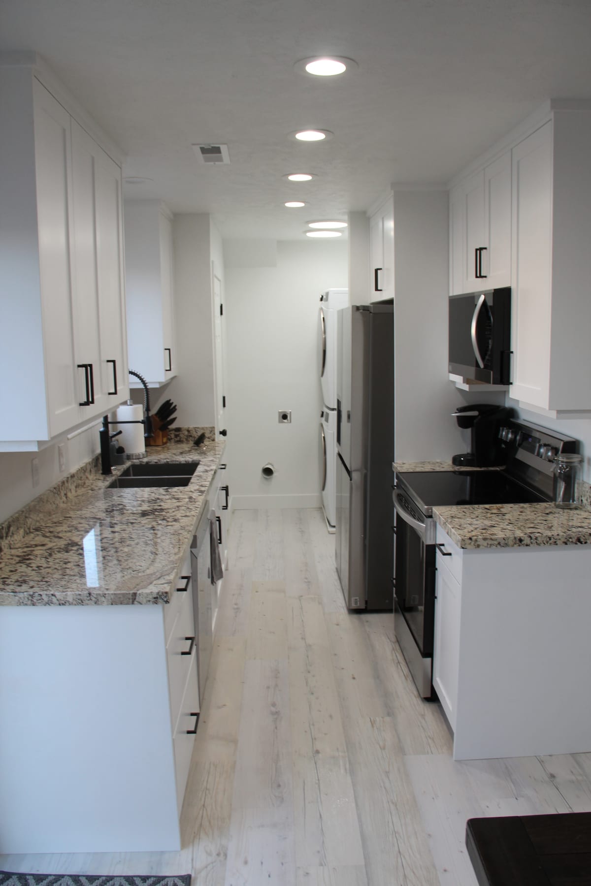 A modern kitchen features granite countertops and white cabinetry, providing a clean and spacious look. Stainless steel appliances, including a refrigerator and oven, are neatly integrated. The flooring is light-colored and extends down the narrow corridor, illuminated by recessed ceiling lights.