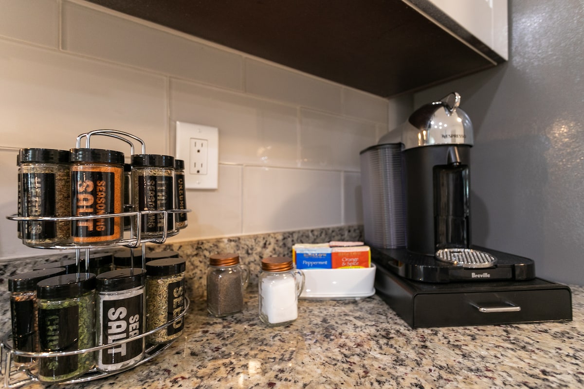 The kitchen countertop showcases a variety of spice jars arranged on a rack, alongside containers filled with sugar and a selection of teabags. A sleek coffee maker is positioned nearby, complemented by a supply of disposable coffee cups.