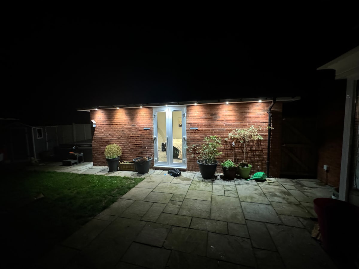 The exterior of the newly converted space is illuminated at night, highlighting its brick façade. Glass patio doors are centered, flanked by potted plants. A stone-paved patio extends in front, providing access to the garden area. The surrounding landscape is softly lit, enhancing visibility.