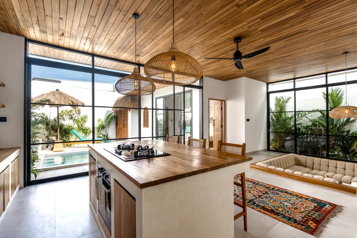 A modern kitchen area features a wooden island with a gas stovetop, surrounded by natural wood accents. Open spaces are filled with light from large glass doors leading to an outdoor area, showcasing a swimming pool and lush tropical greenery. A cozy seating area adds to the inviting ambiance.