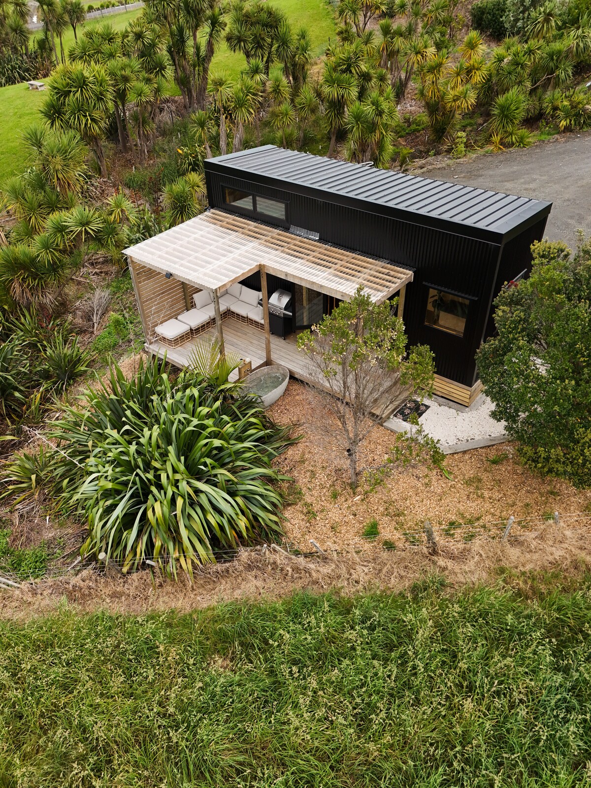 The tiny home is nestled among lush greenery, featuring a covered outdoor area and a deck that showcases a mix of native plantings. The structure's modern black exterior contrasts with the surrounding landscape, creating a serene and inviting retreat.