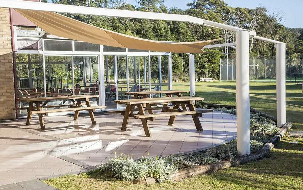 An outdoor seating area is highlighted, featuring several wooden picnic tables arranged on a tiled surface. A shade sail provides coverage above, and lush greenery borders the space, enhancing the open environment. The background includes a grassy area and a tennis court.