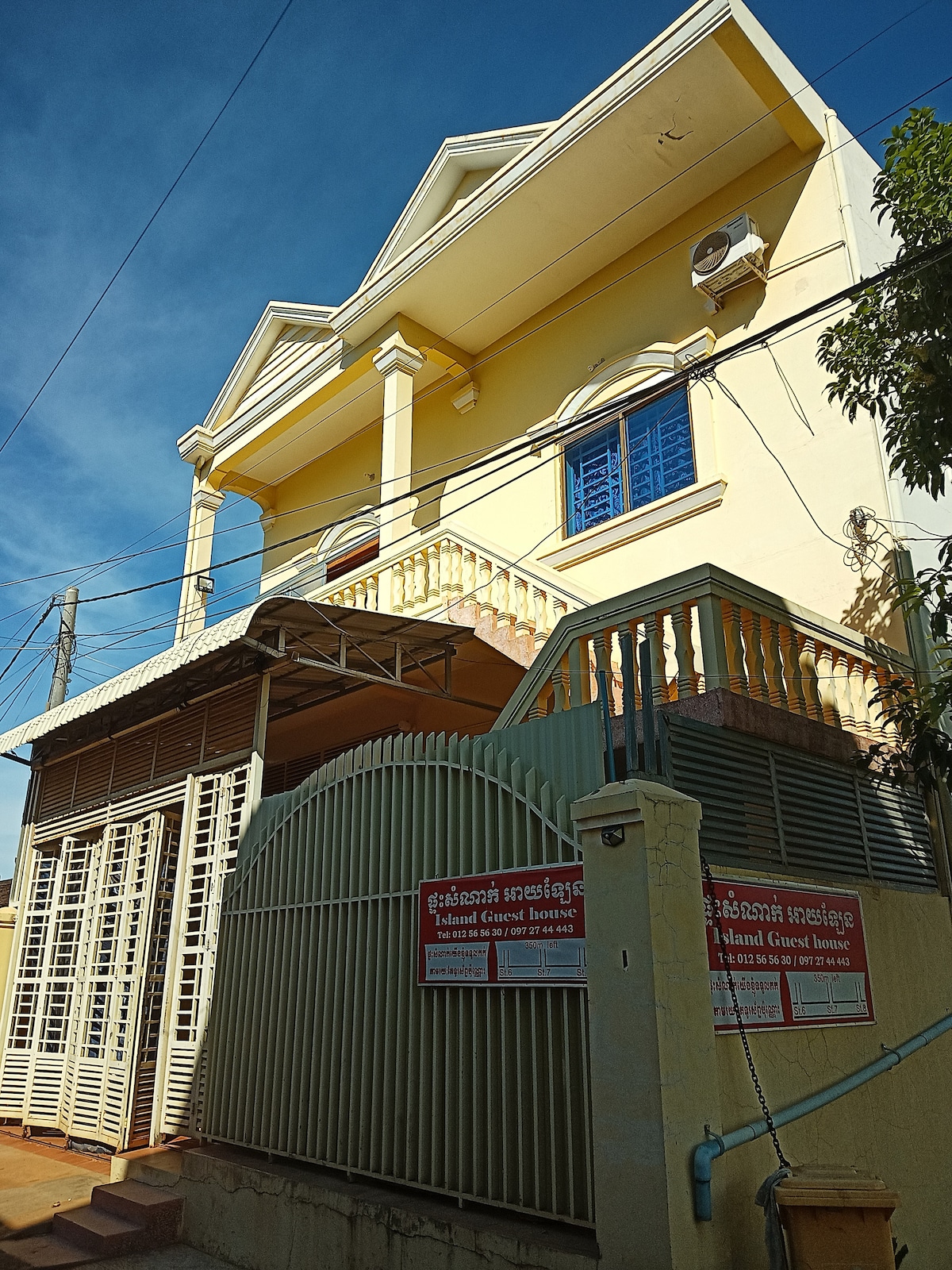 The exterior of a two-story yellow guesthouse is visible, featuring a gated entrance. A stairway leads to a balcony with white railings. Blue windows adorn the upper level, while the ground floor is accessed through a covered porch area with protective metal grilles.