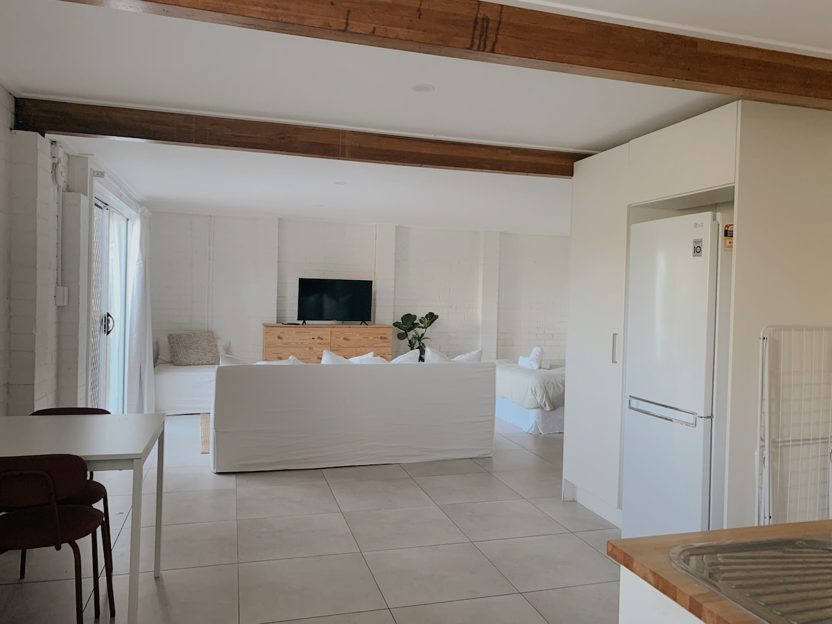 A spacious interior is presented, featuring a light-filled living area with a comfortable sofa and a wall-mounted television. A dining area with a table and chairs is visible, alongside a modern kitchen that includes a refrigerator. Soft textiles add warmth to the space.
