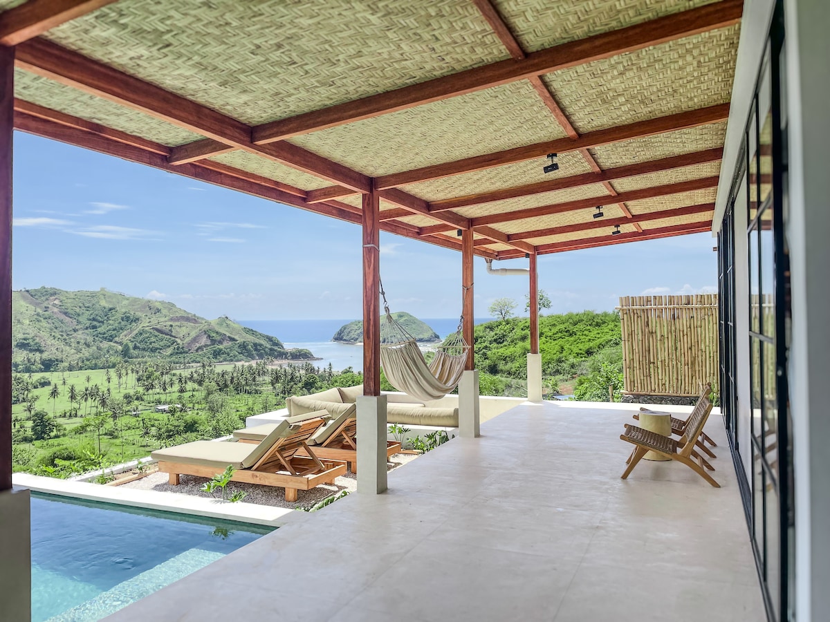 An outdoor terrace is shown, featuring a sunken lounge area and lounge chairs overlooking a scenic view of the ocean and greenery. A hammock is hung between two wooden beams, providing a relaxing space. The infinity pool reflects the surrounding landscape.