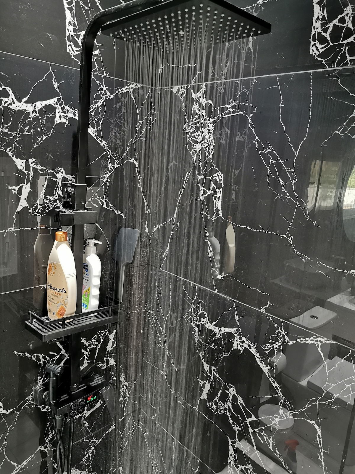 A modern shower setup is showcased with a rainfall showerhead positioned above. Black marble walls create a sleek and luxurious backdrop. Various toiletries are neatly arranged on a wall-mounted shelf, enhancing convenience and functionality.