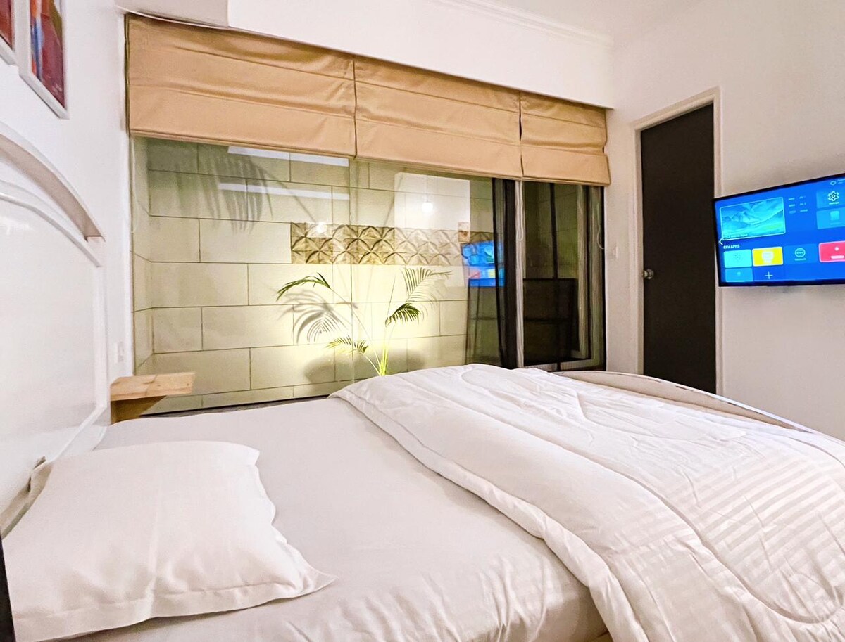 The sleeping area features a spacious bed with white linens and pillows. A large window is dressed with a beige shade, allowing natural light to illuminate the interior. An accent wall displays plant motifs, and a flat-screen TV is mounted nearby.