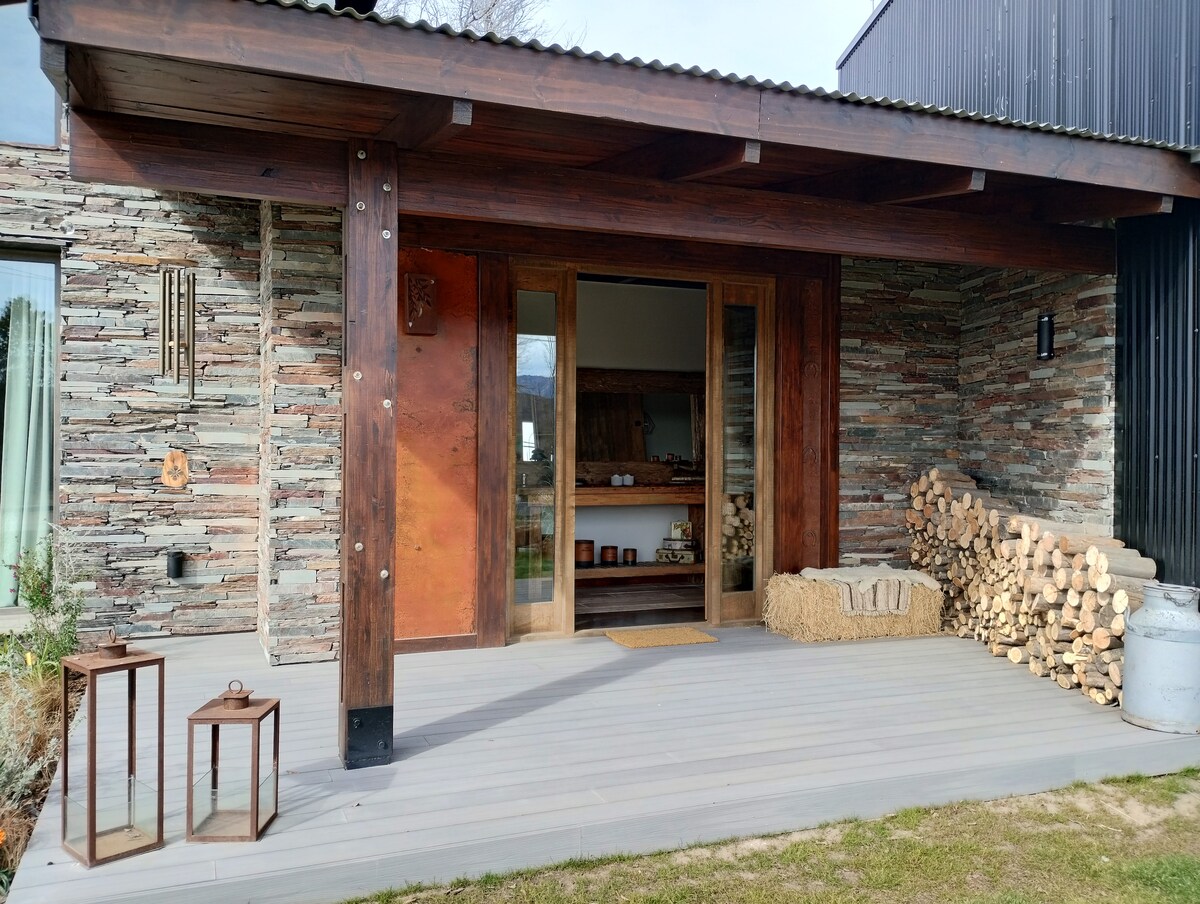 The exterior features a spacious wooden deck with two side tables and neatly stacked firewood. Large wooden doors, framed by stone walls, lead into the inviting interior. Natural light filters through the surrounding openings, creating a welcoming entrance.