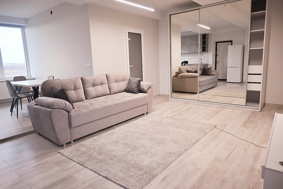 A spacious living area features a soft gray sofa facing a large mirror that reflects the room's design elements. A light-colored area rug is positioned on the wooden floor, while a dining table and chairs are visible in the background, enhancing the room's functionality.