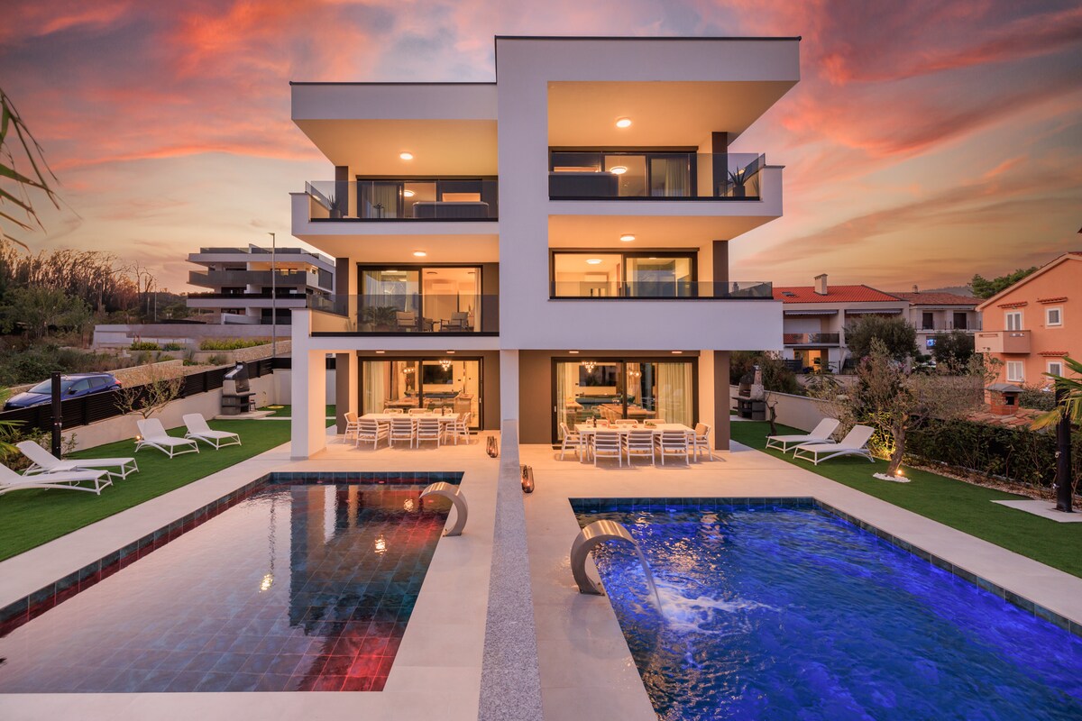 A modern two-story villa is situated amidst landscaped gardens, featuring a heated pool and jacuzzi on either side. Outdoor seating areas are arranged for relaxation, while large glass doors provide a view of the interior. A warm sunset sky enhances the setting.