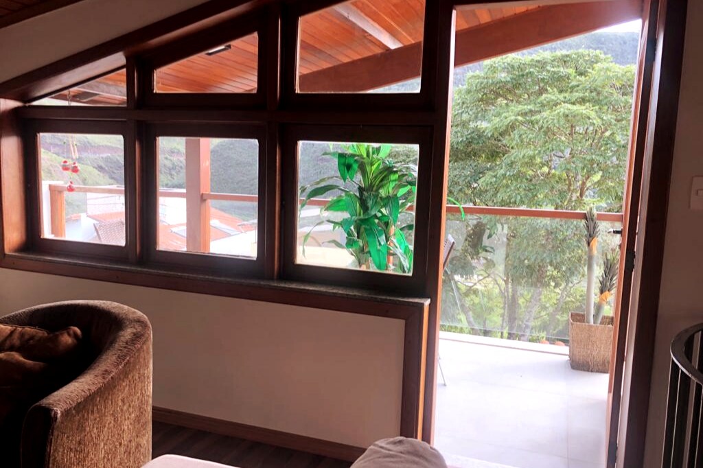 A spacious interior is highlighted by large windows that frame a serene view of the surrounding greenery and outdoor balcony. Natural light fills the room, enhancing the warm wooden tones of the ceiling and the cozy seating area.