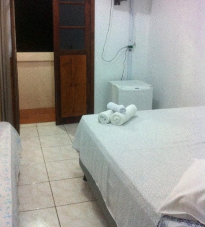 The room features two beds with crisp white linens. A small refrigerator is positioned nearby, along with neatly rolled towels placed on the bed. A wooden door provides access to a balcony, while the tiled floor adds a clean, simple aesthetic.