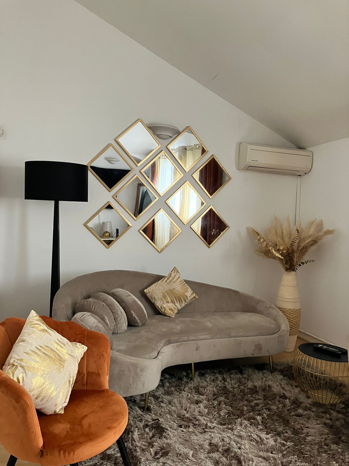 A cozy living area features a curved grey sofa accented with decorative cushions and an orange chair. A decorative vase with dried plants is positioned beside a textured table. A wall of diamond-shaped mirrors enhances the space, complemented by soft lighting from a black floor lamp.
