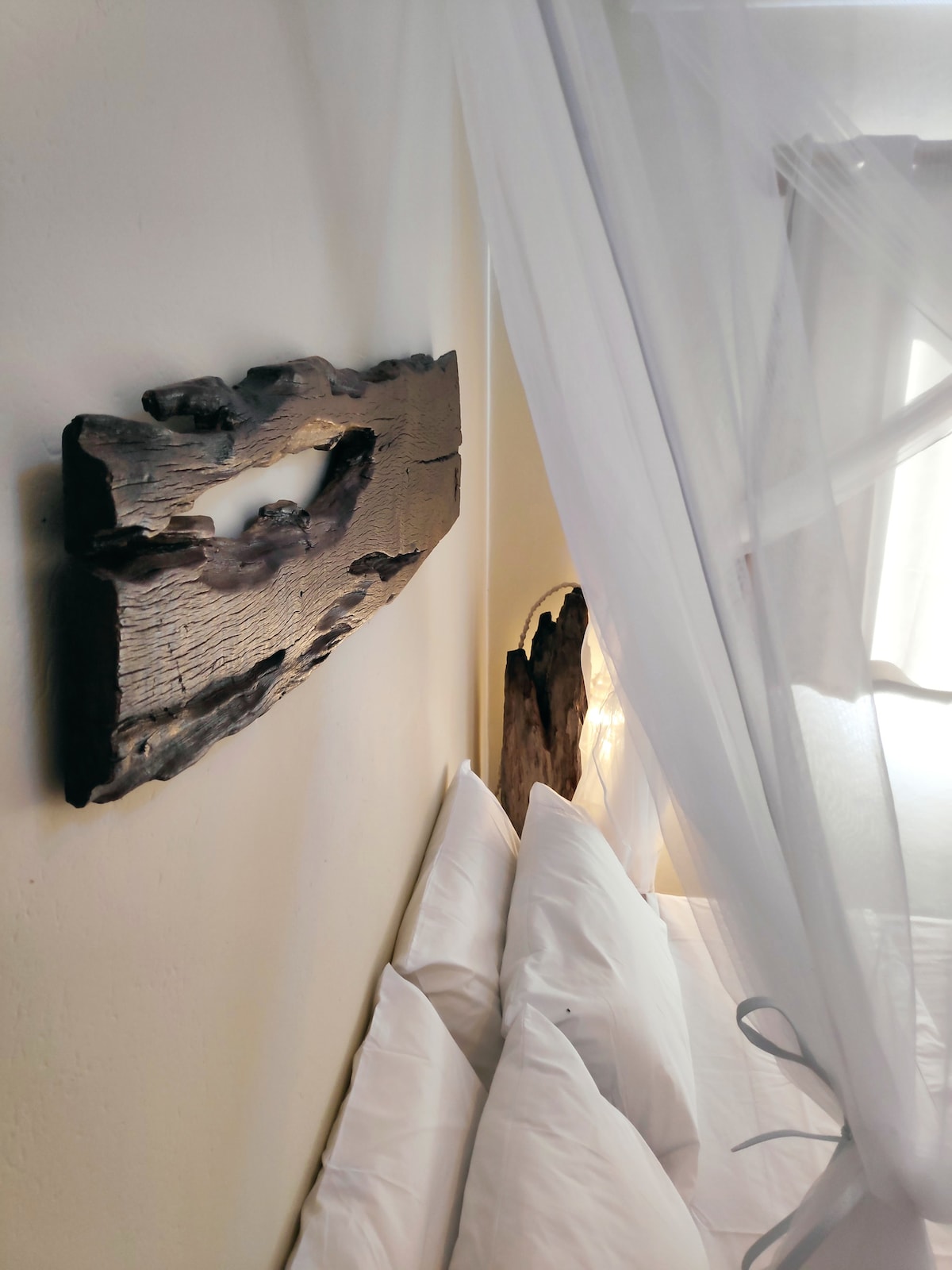 A serene sleeping area features a spacious bed adorned with plush, white pillows. Above the bed, unique wooden wall art adds character, while sheer white drapery softly frames the space, allowing light to filter in gently.