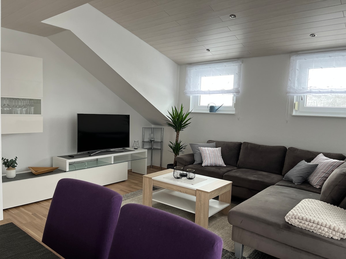 A spacious living area features a comfortable sectional sofa and a modern coffee table. A television is placed on a sleek entertainment unit. Natural light enters through two windows, complemented by curtains. A decorative plant adds a touch of greenery to the inviting space.