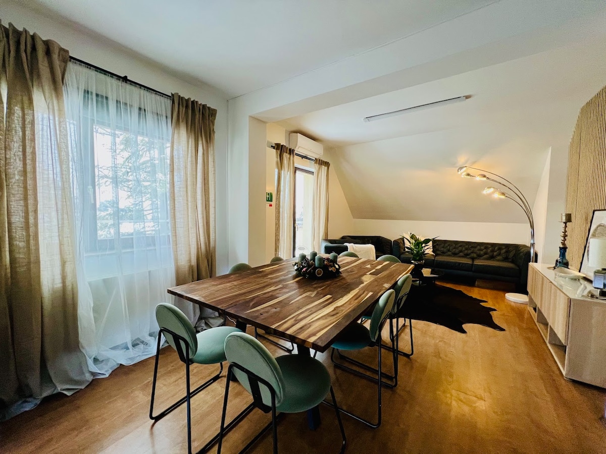 A spacious dining area features a large wooden table surrounded by green chairs. Natural light streams through large windows adorned with sheer curtains. A comfortable seating area is visible in the background, highlighted by dark furniture and decorative plants.
