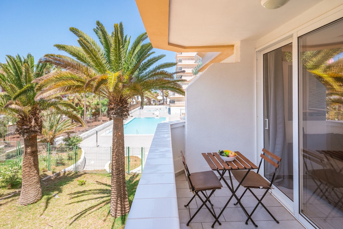 An outdoor terrace is presented, featuring a table with two chairs and a small fruit arrangement. Lush palm trees frame the view, while a swimming pool is visible in the background, contributing to a relaxed outdoor setting.