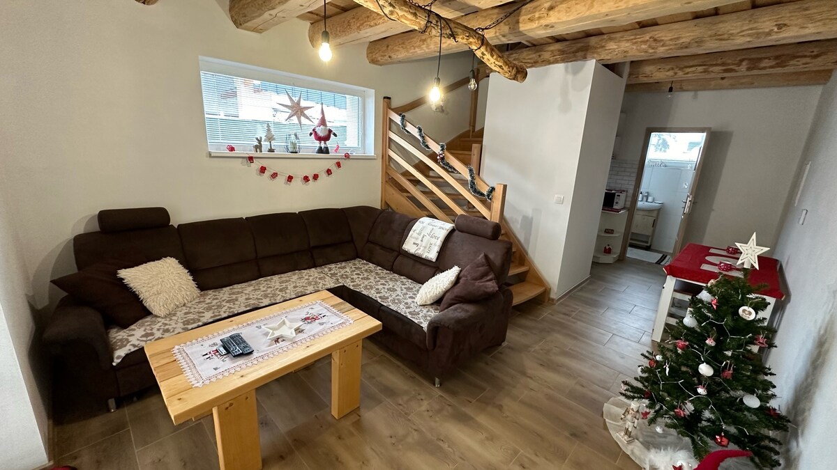 A spacious living area features a large, dark brown sectional sofa accompanied by a wooden coffee table. A small Christmas tree decorates the corner. Visible wooden beams and a staircase lead to an upper level, while a cozy ambiance is created by soft lighting.