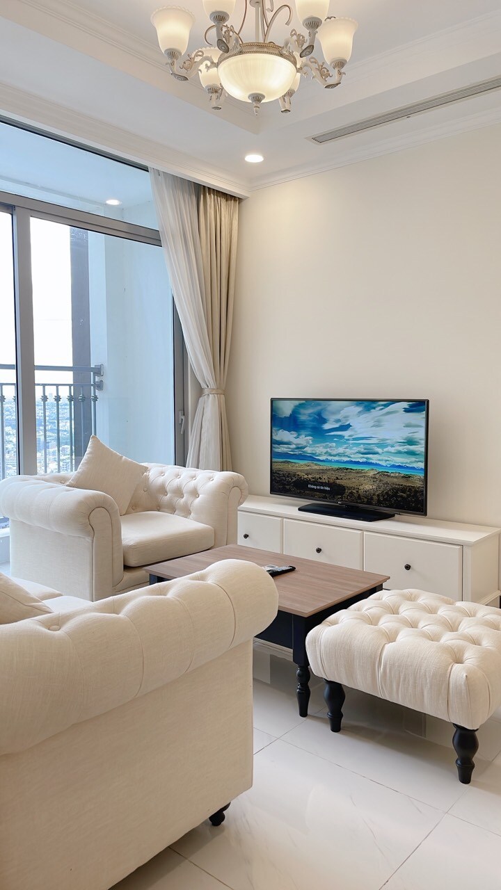 A light-filled living area features two plush armchairs arranged beside a wooden coffee table. A neutral-toned sofa is positioned opposite a console with a flat-screen television displaying a scenic landscape. Floor-to-ceiling windows allow natural light to enhance the space.