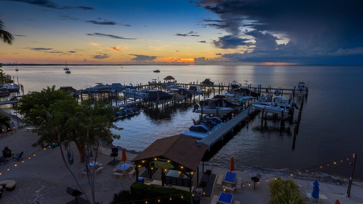 The image captures a tranquil waterfront scene at sunset, with boats docked at a marina. The sky is painted in soft shades of orange and purple, reflecting on the water's surface. Dimly lit outdoor seating and beach umbrellas are visible along the shore, creating a serene atmosphere.