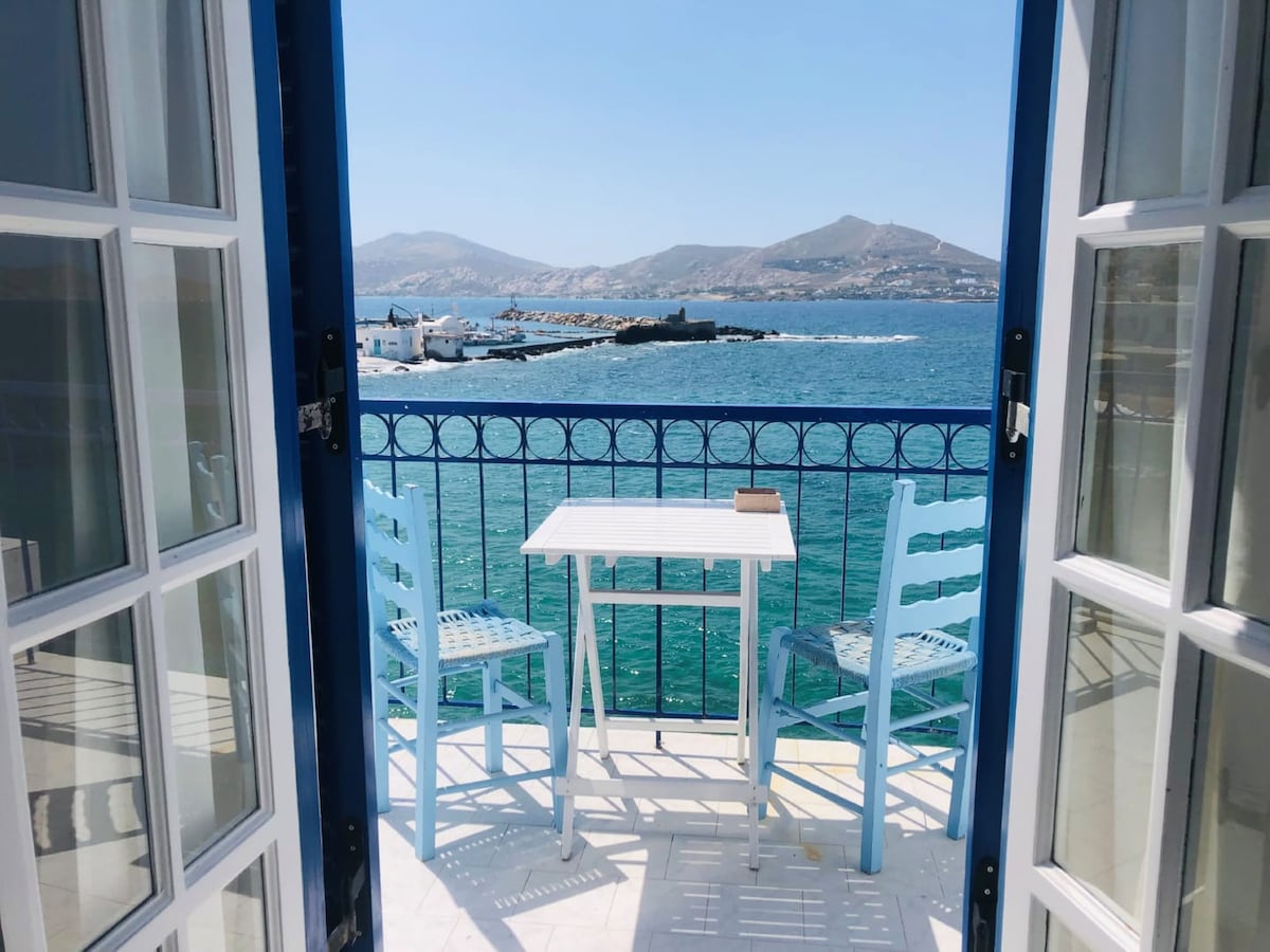 A private balcony offers a clear view of the sparkling sea and distant hills. Light blue outdoor furniture, including a table and two chairs, is arranged invitingly under bright sunlight. The balcony rail features decorative ironwork, enhancing the scenic coastal ambiance.