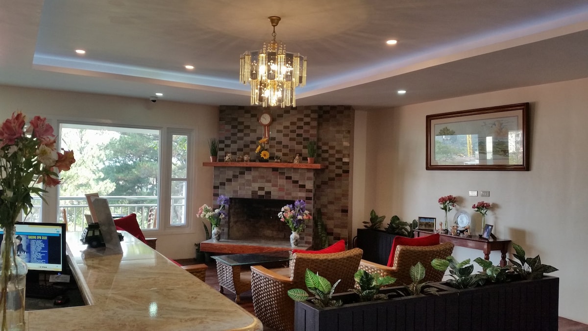 A welcoming living area presents a cozy seating arrangement with wicker chairs and vibrant cushions. A stone fireplace is the room's focal point, complemented by a decorative clock and fresh flowers. Large windows allow natural light and offer views of the surrounding greenery.