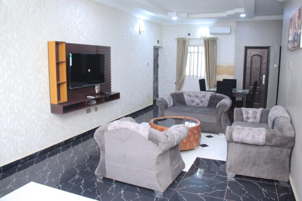 A modern living area is furnished with two plush sofas and a circular coffee table. A wall-mounted television is positioned near a window with drapes. Light fixtures are installed in the ceiling, enhancing the bright atmosphere of the room.