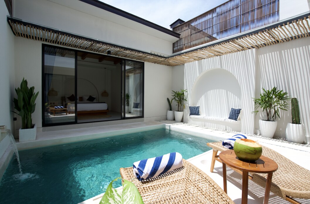 An outdoor area features a refreshing pool surrounded by elegant loungers and a small table. Potted plants add greenery to the space, while large sliding glass doors provide a view into the inviting interior, which is illuminated by natural light.