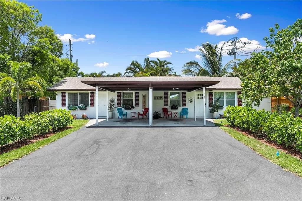 A welcoming exterior view features a single-story apartment with a covered porch. Comfortable seating is arranged in vibrant colors. Lush greenery lines the driveway, providing a serene atmosphere. Tropical trees and blue skies enhance the natural surroundings, highlighted by sunny weather.