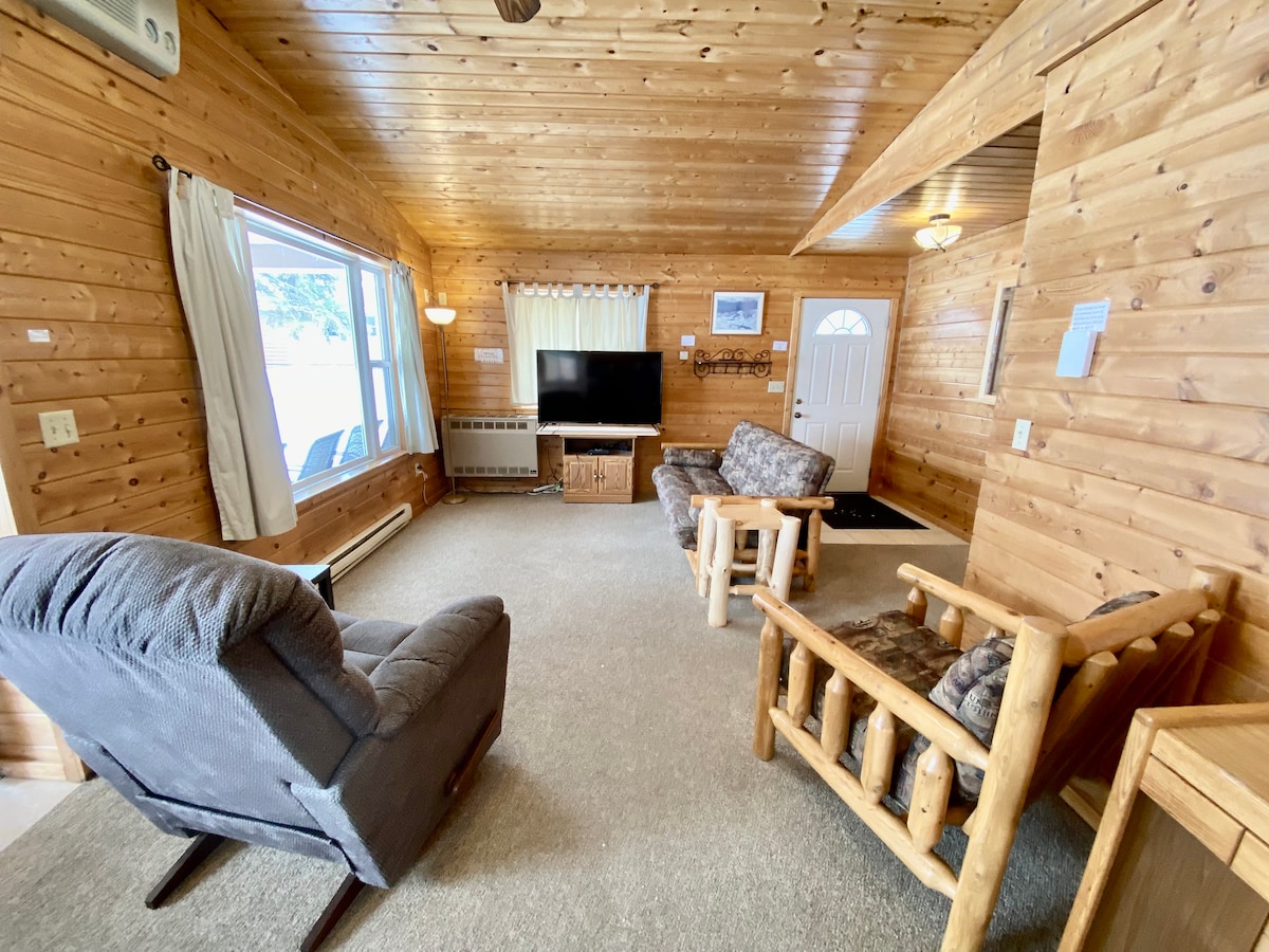 A welcoming living area features wooden walls and a carpeted floor. A large television is placed against one wall, surrounded by comfortable seating arrangements, including a recliner and a rustic wooden chair. Natural light enters through large windows, enhancing the cozy ambiance.