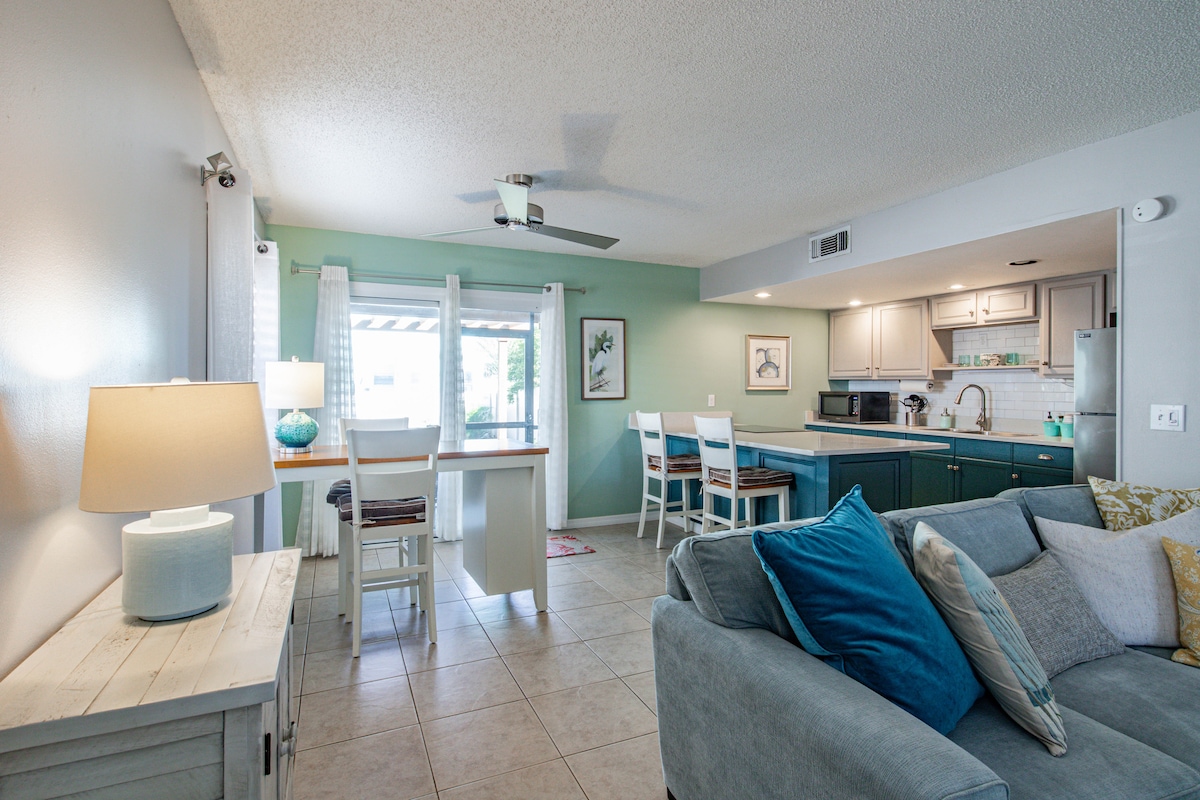 An open-concept living area features a comfortable sofa adorned with blue and yellow pillows. A dining table for four sits adjacent to the modern kitchen, illuminated by natural light from the sliding glass doors leading to the outside. Light tile flooring contributes to the airy feel.