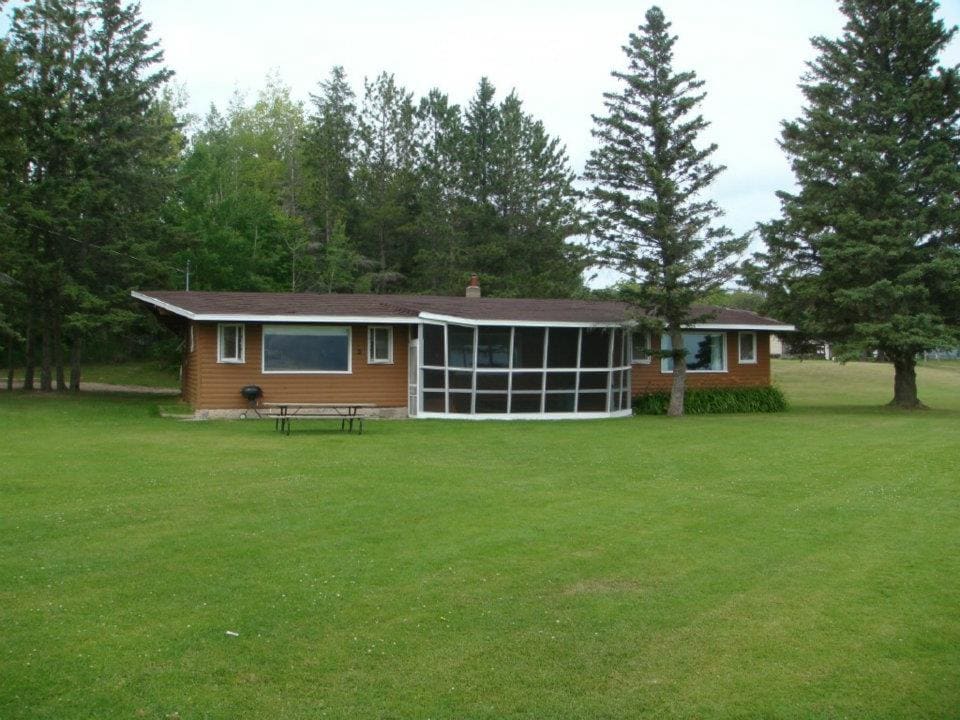 A spacious cabin exterior is framed by tall green trees and a well-maintained lawn. The structure features a screened porch and large windows that provide ample natural light. A charcoal grill is visible on the patio, alongside a table for outdoor dining.