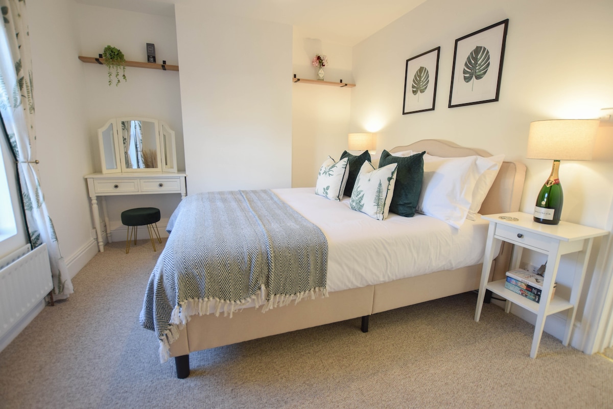 A stylish bedroom features a spacious bed dressed with white linens and accented by green throw pillows. A dressing table with a mirror is positioned against the wall, alongside a small stool. Soft lighting is provided by two bedside lamps, creating a calming ambiance.