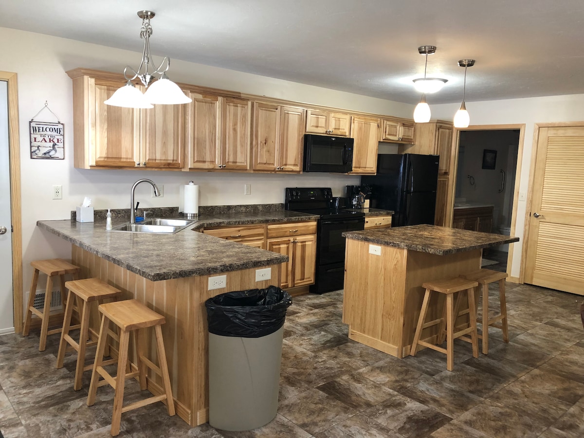 An open kitchen features wooden cabinetry and a dark countertop, complemented by stainless steel appliances. Two kitchen islands with seating for four are visible, along with hanging light fixtures above. A welcomed sign decorates one wall, adding character to the functional space.