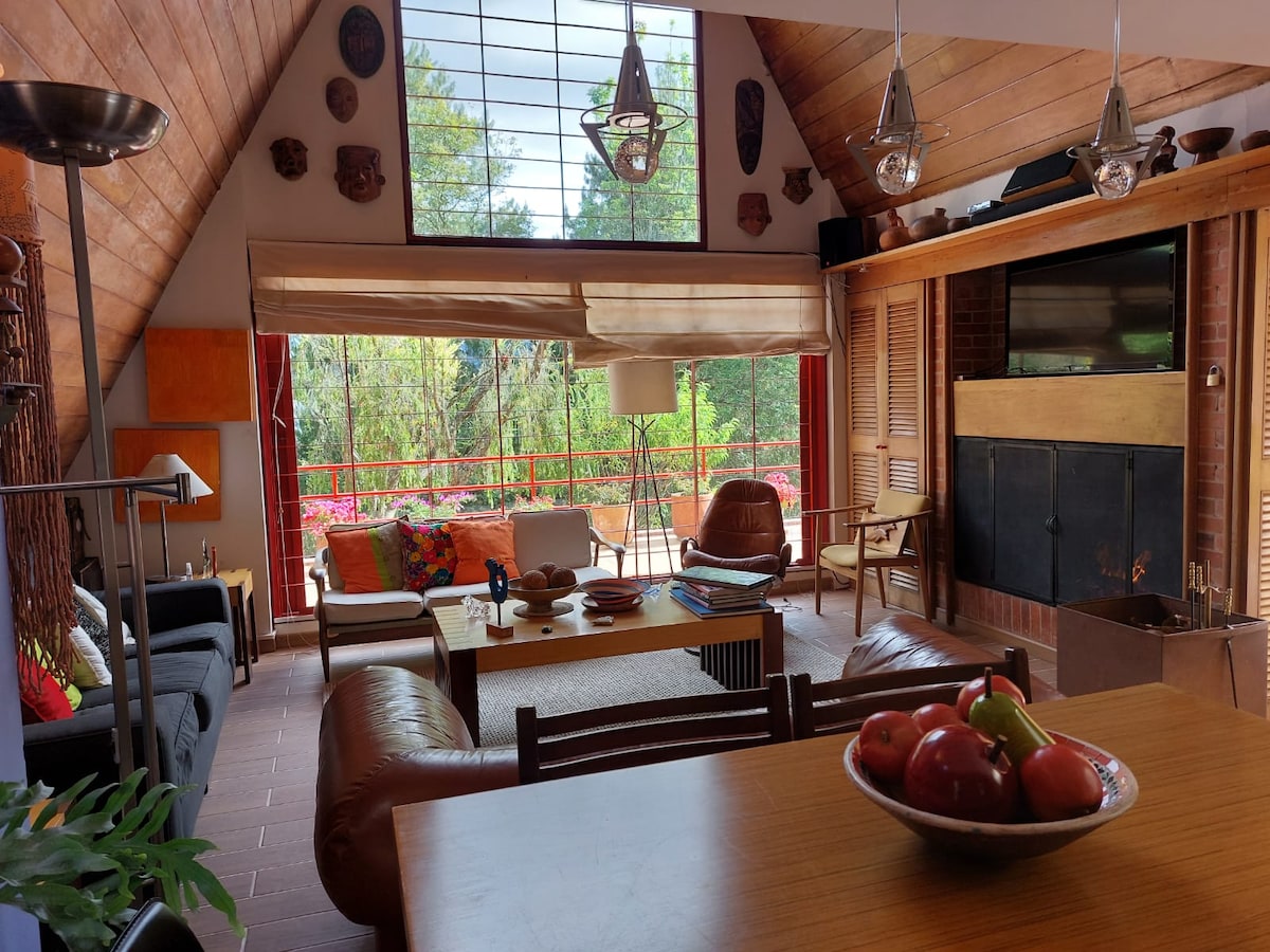 The living space features high vaulted wooden ceilings with large windows offering views of the surrounding greenery. A mix of comfortable seating arrangements is highlighted, including a brown leather sofa and a wooden coffee table. A fruit bowl sits on a dining table, enhancing the welcoming atmosphere.