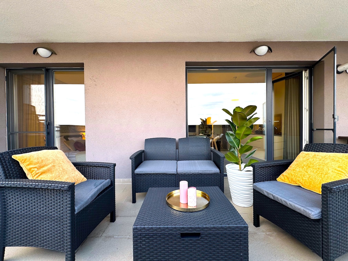 A cozy balcony features a seating arrangement with two dark wicker chairs and a matching couch adorned with yellow cushions. A small circular table sits in the center, complemented by a decorative plant in a white pot. Distant city views are visible through the open doors.