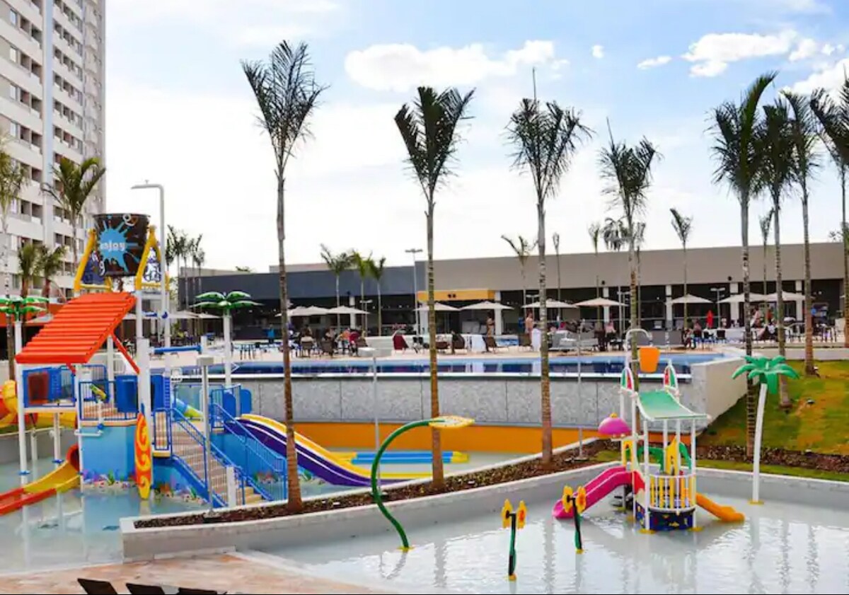 A family-friendly water play area features colorful slides and play structures surrounded by palm trees. Shallow pools with waterslides offer a vibrant atmosphere. Lounging areas are visible in the background, providing ample seating options for relaxation.