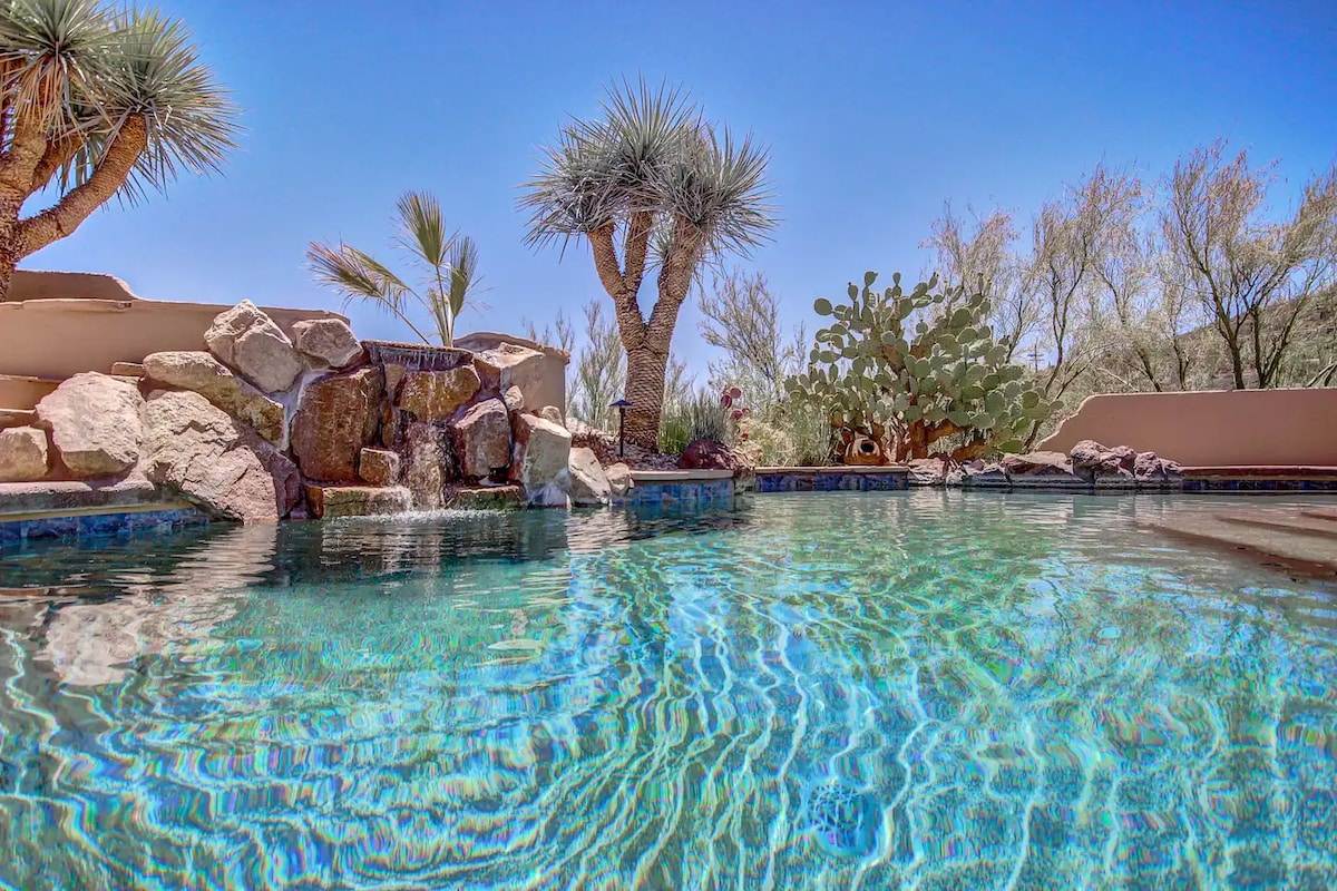 A tranquil swimming pool is surrounded by natural landscaping, featuring cacti and palm trees. The water reflects the clear sky above, while a rock formation adds a decorative waterfall element. This serene setting provides a relaxing atmosphere.