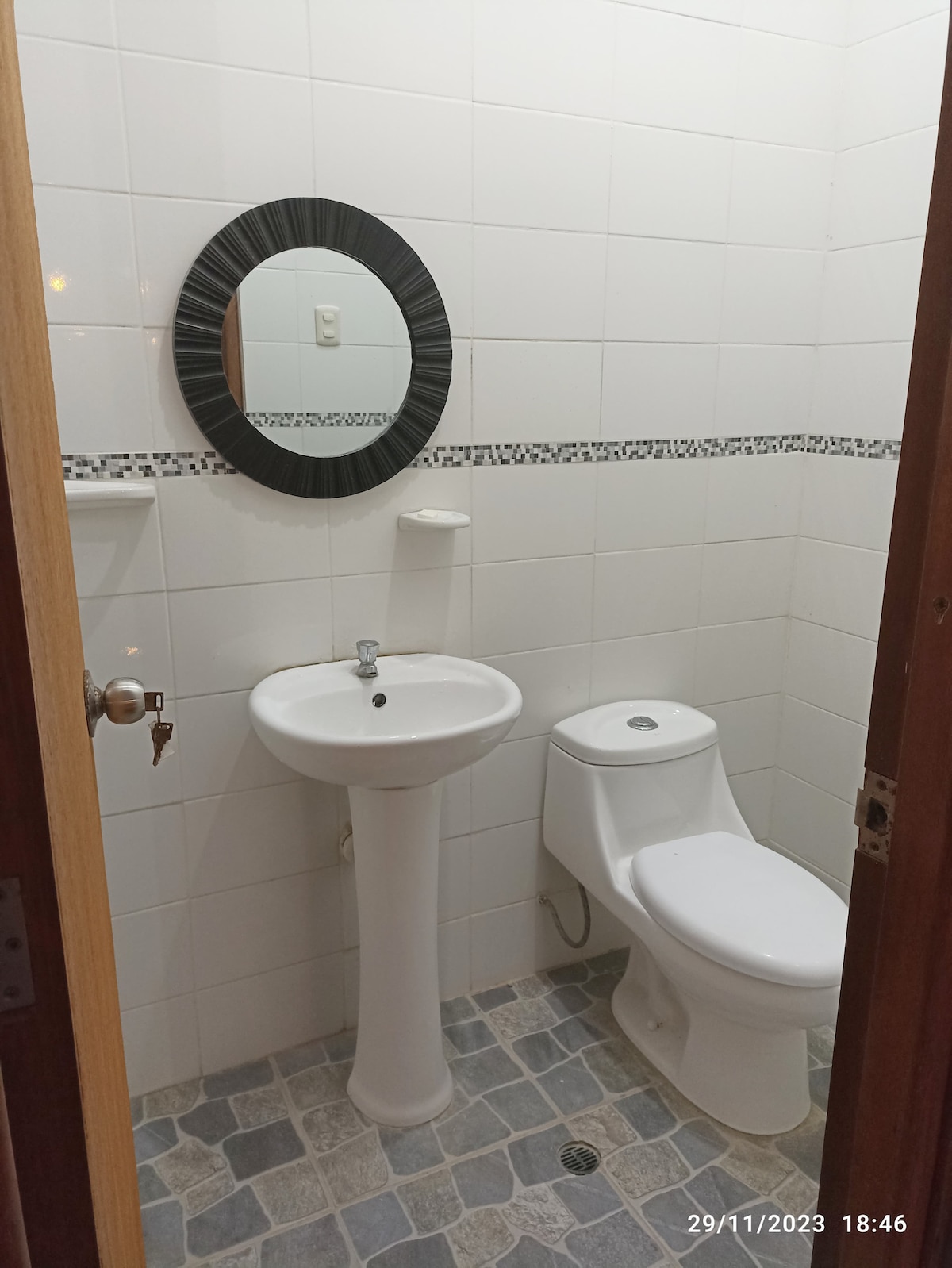 A well-lit bathroom is displayed, featuring white tiled walls and a light gray floor. A round mirror with a black frame hangs above a white sink, while a white toilet is positioned nearby. Basic amenities are provided, ensuring functionality and convenience.