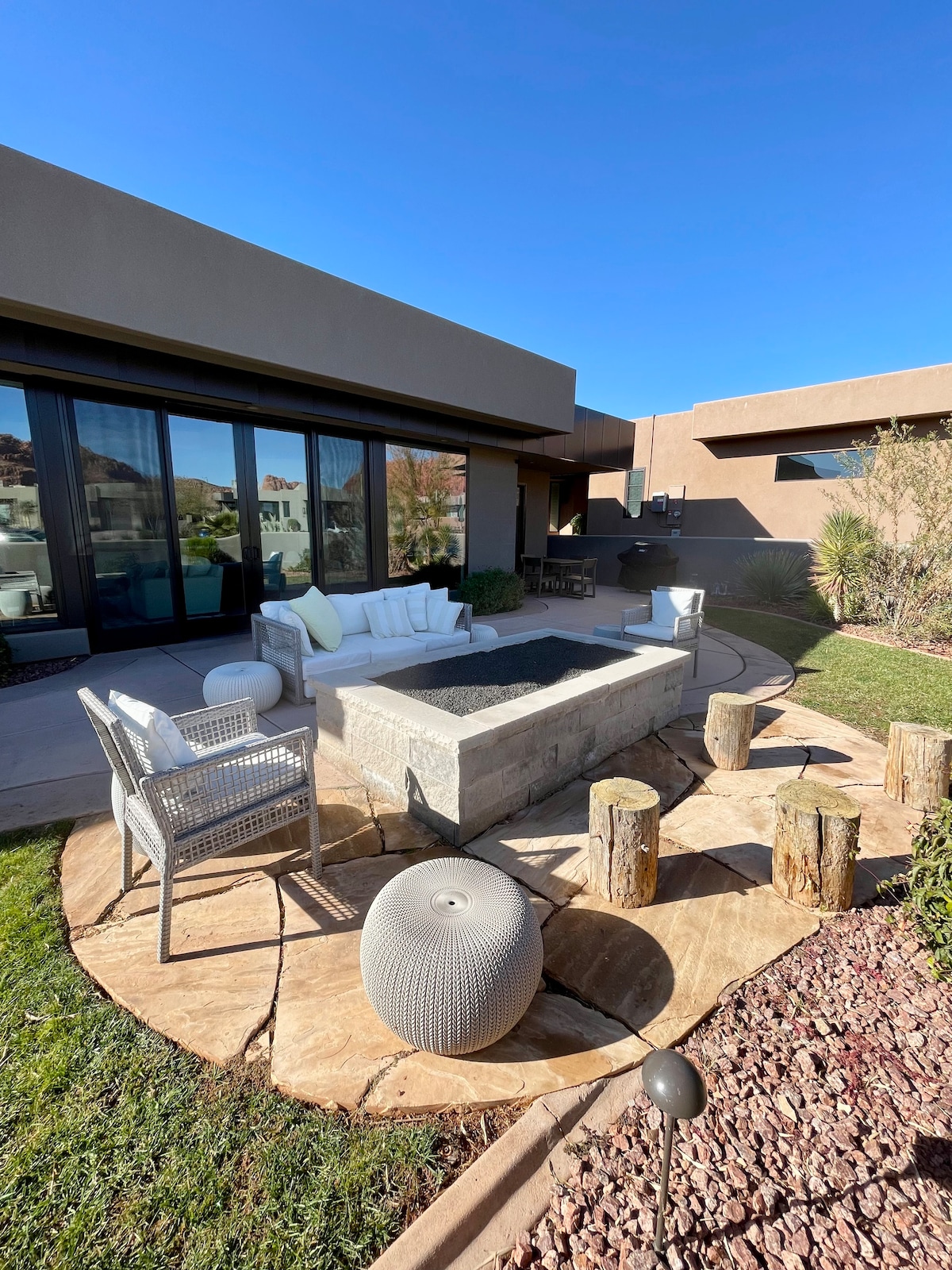 An outdoor seating area features a white sofa and woven chairs surrounding a stone fire pit, with circular seating made from tree stumps. A pathway of large stone slabs leads to a well-maintained lawn, framed by colorful landscaping and clear blue skies.