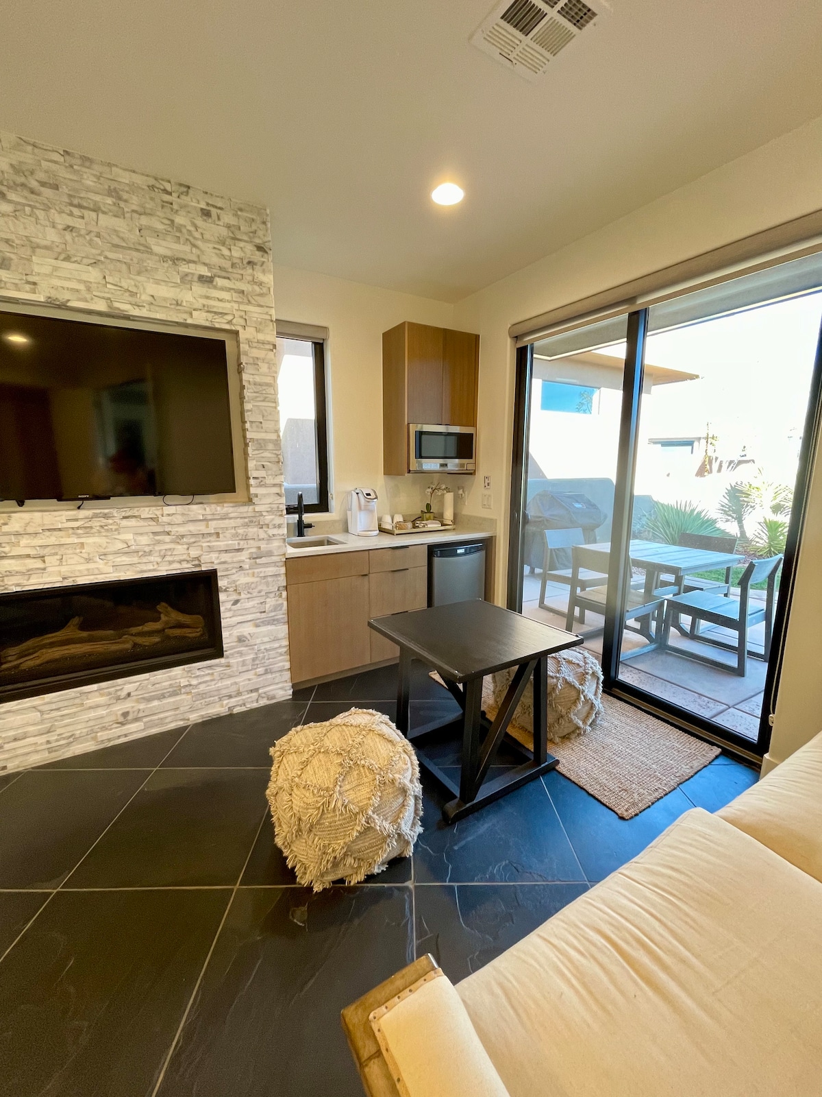 A modern living area features a flat-screen TV mounted above a sleek fireplace, complemented by a kitchen equipped with essential appliances. Natural light filters through large glass doors, leading to an outdoor seating area, while textured poufs add a cozy touch to the space.
