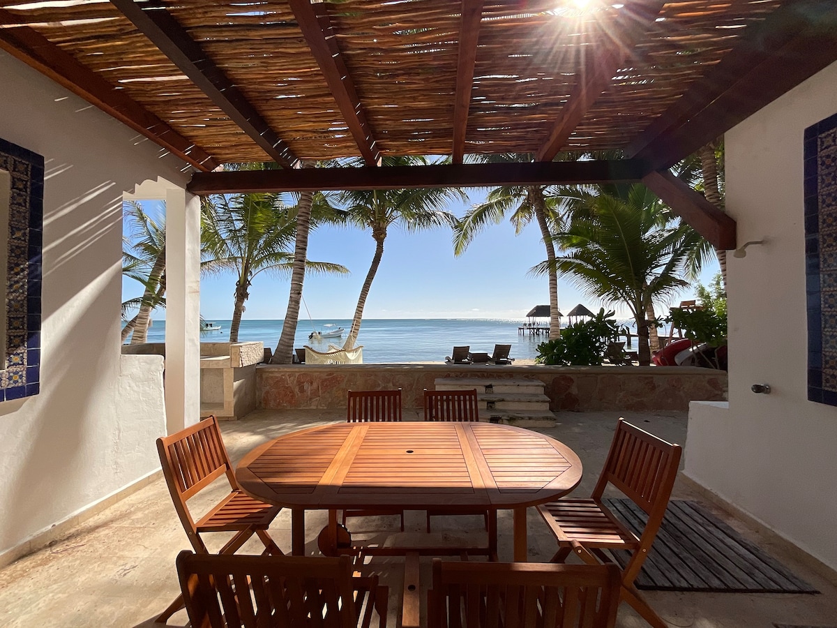 An outdoor dining area is featured under a shaded pergola, showcasing a round wooden table surrounded by eight chairs. Palm trees sway nearby, with a view of the ocean and a beach in the distance, creating a serene setting for meals.