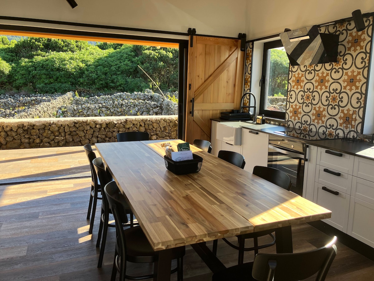 A spacious kitchen area features a large wooden dining table surrounded by black chairs. Floor-to-ceiling glass doors open to reveal views of lush greenery and stone walls, while patterned wallpaper adds a decorative touch to the room.