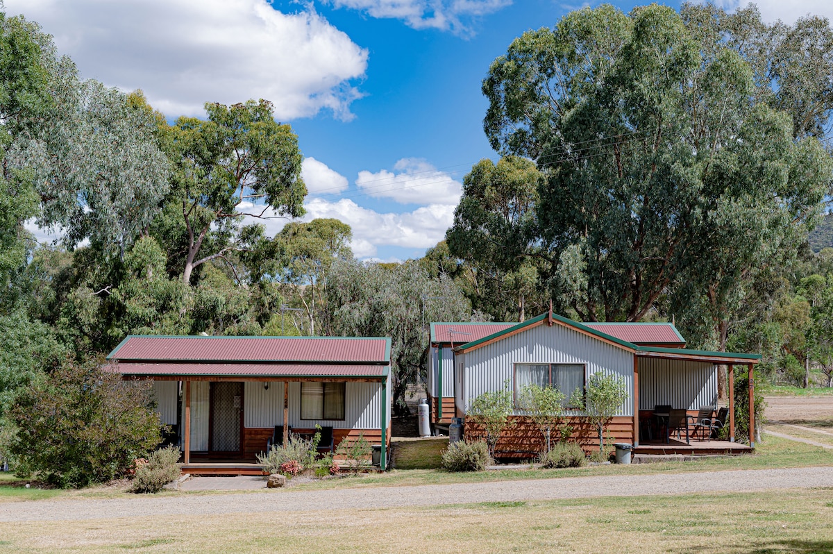 The image features two deluxe cabins set amidst lush greenery. Each cabin has a distinct design, with a front porch and large windows. The surrounding landscape includes rolling hills and mature trees under a partly cloudy sky, creating a serene outdoor environment.