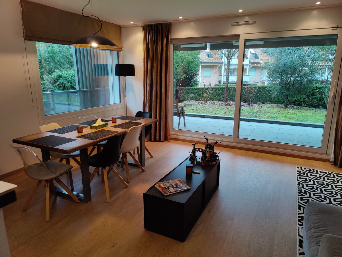 A modern dining area features a wooden table surrounded by six chairs. Floor-to-ceiling windows provide a view of the garden outside, while natural light illuminates the space. A stylish black coffee table with magazines adds to the room's functional design.
